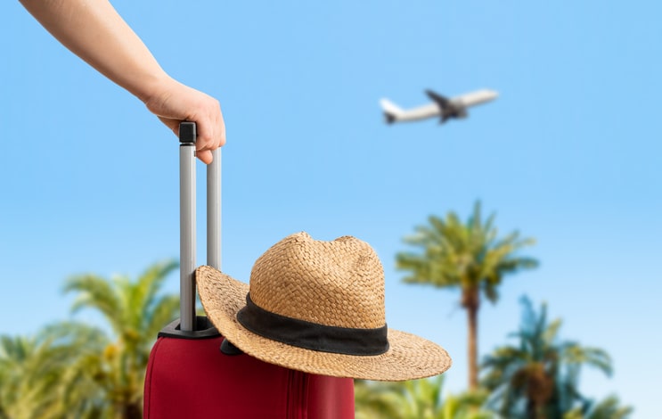 A hand pulling a suitcase with a hat, with palm trees and an airplane in the background